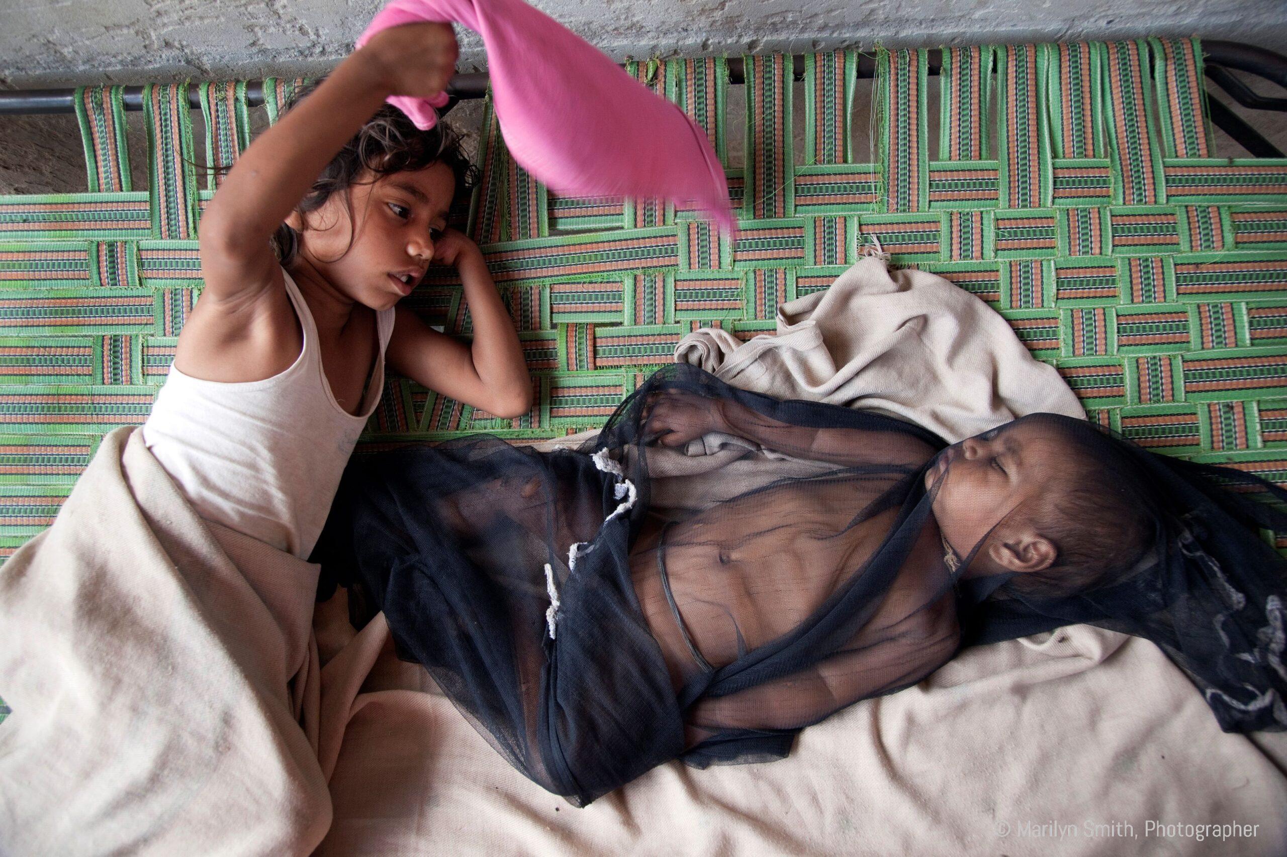 A young girl swatting flies while her baby brother sleeps.